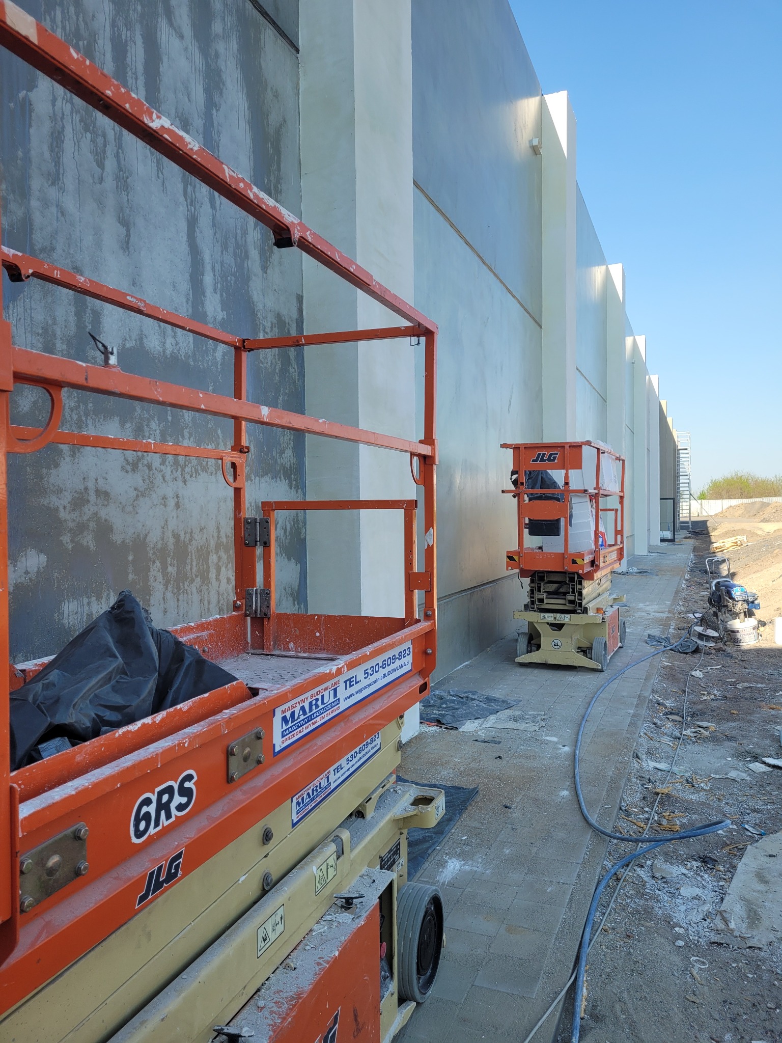 Dwie pomarańczowe platformy podnośnikowe JLG stojące obok ściany budynku w trakcie prac wykończeniowych, z widocznymi śladami świeżo nałożonej warstwy szarej substancji, prawdopodobnie gładzi...