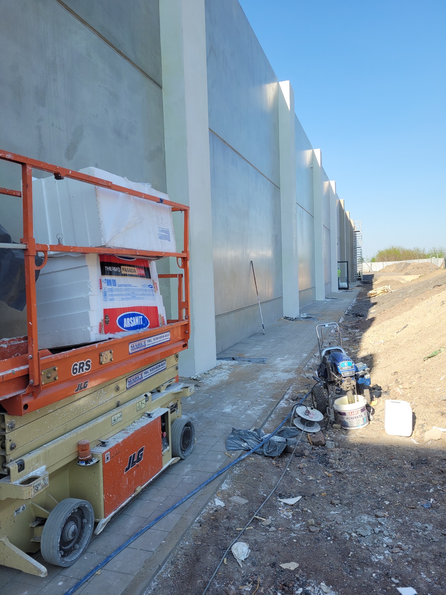 Świeżo pomalowana elewacja budynku przemysłowego z widocznym podnośnikiem koszowym z materiałami izolacyjnymi Arsanit Thermo Fasada Extra, agregatem malarskim Texspray i wiadrem farby Kabe Perfekta.