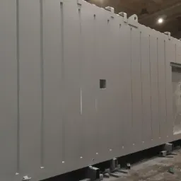 Świeżo pomalowany na szaro kontener z widocznym otworem okiennym zabezpieczonym folią, umieszczony na metalowych wspornikach w hali produkcyjnej, obok widoczny kompresor i drabina.