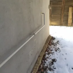 Szara rura gazowa zamontowana na szarej ścianie budynku, widoczne mocowania, fragment drewnianych drzwi i śnieg na ziemi.