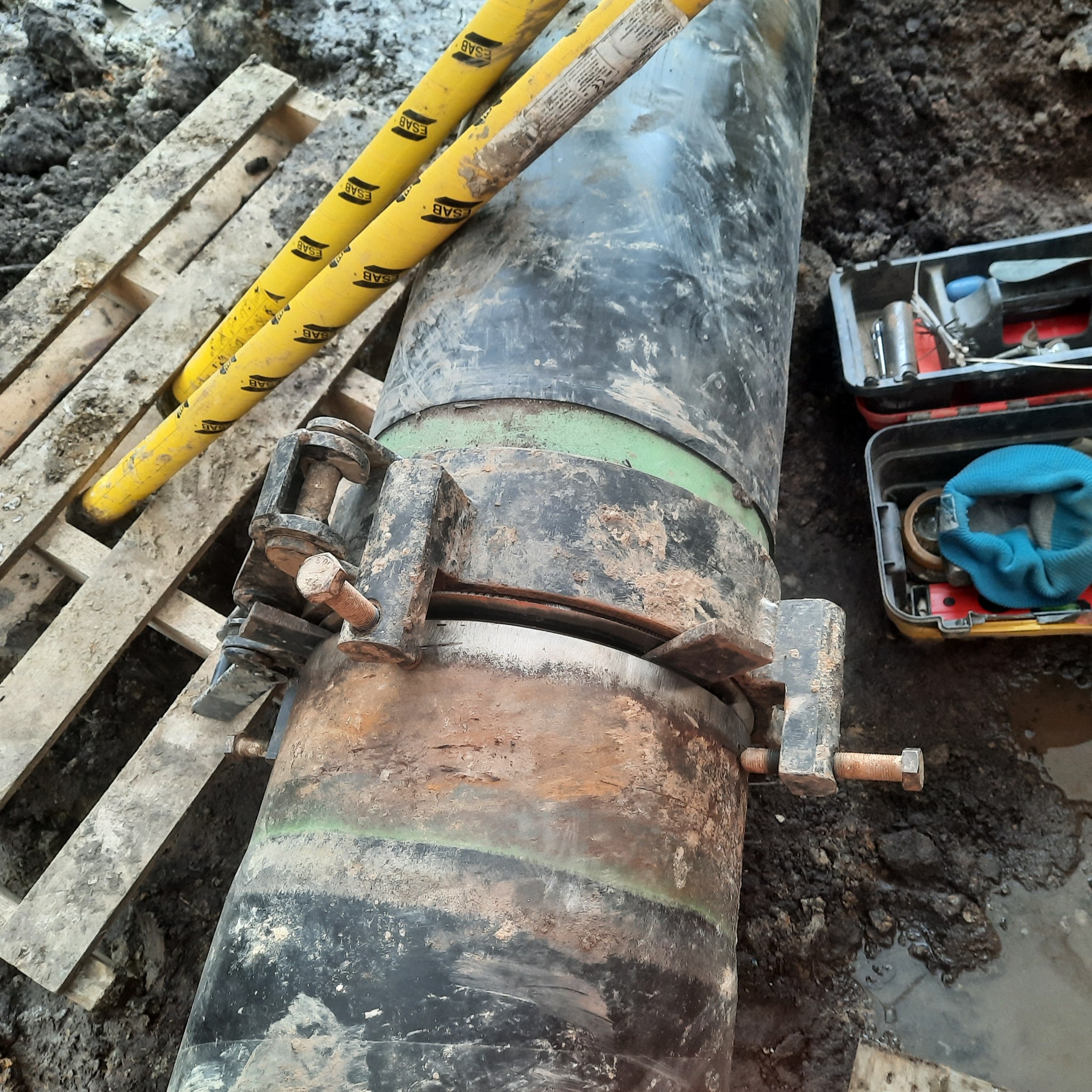 Złącze kołnierzowe na rurociągu gazowym w wykopie, narzędzia w skrzynce obok, widoczne żółte osłony spawacza ESAB.