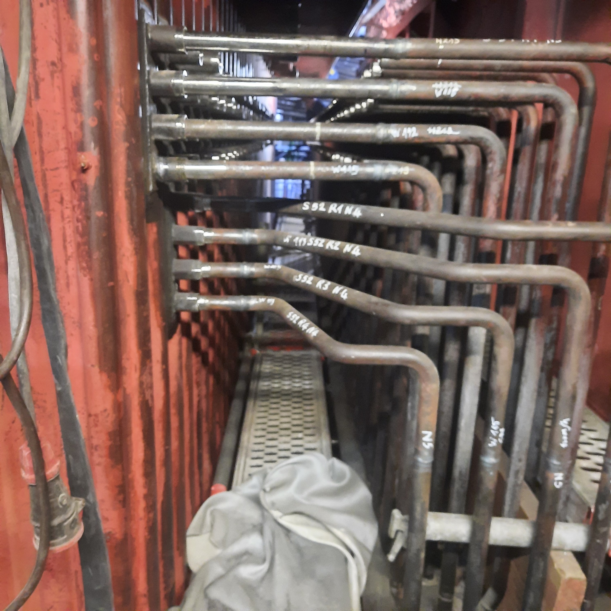 Rząd metalowych rur z widocznymi spawami i oznaczeniami, część instalacji wewnątrz konstrukcji stalowej, z widocznymi przewodami elektrycznymi i odzieżą roboczą w tle.