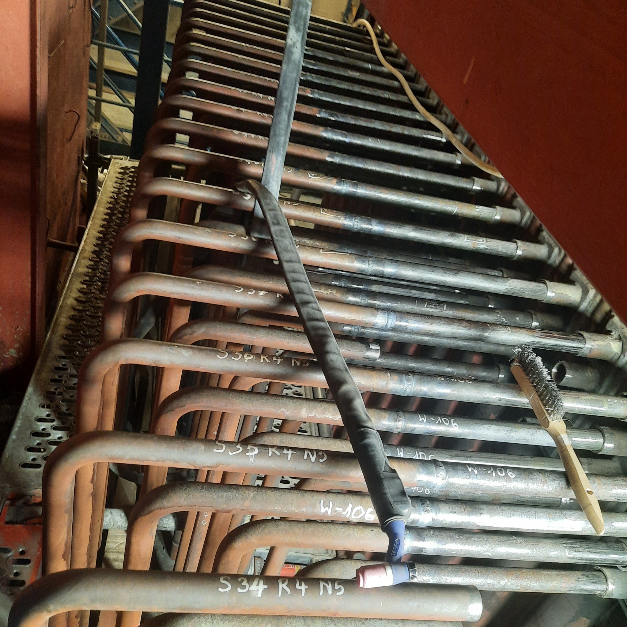 Rzędy metalowych rur z oznaczeniami, częściowo pomalowane, z widocznym przewodem spawalniczym i szczotką drucianą; instalacja przemysłowa w Jaśle.