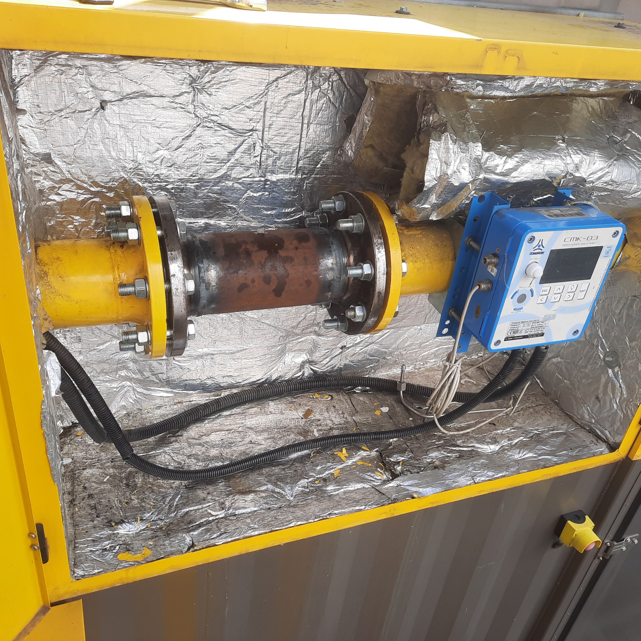 Żółta rura gazowa z zamontowanym miernikiem przepływu gazu CMK-03 w izolowanej obudowie. Widoczne ślady spawania na rurze.