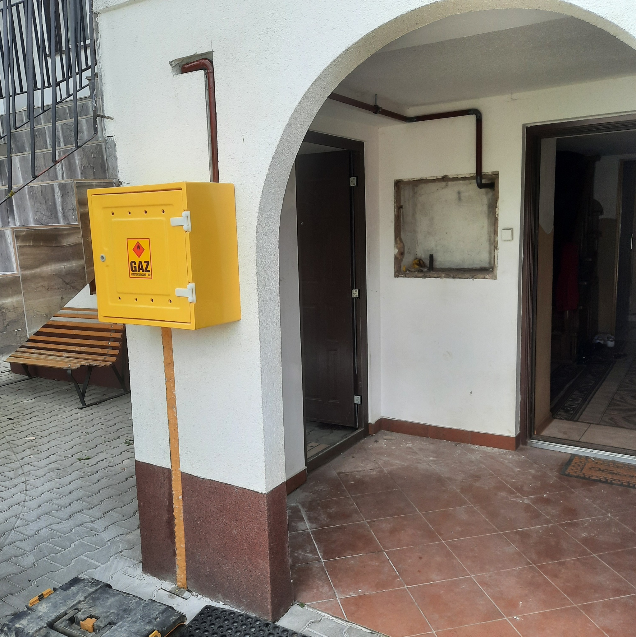 Żółta skrzynka gazowa z napisem 'GAZ' zamontowana na ścianie budynku, widoczne rury gazowe i wejście do budynku z łukowym przejściem.