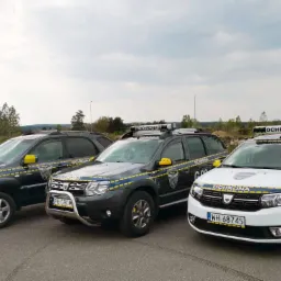 Trzy samochody patrolowe z napisem 'OCHRONA' stojące na parkingu, każdy z innym modelem i kolorem, ale z podobnym żółto-niebieskim oznakowaniem.