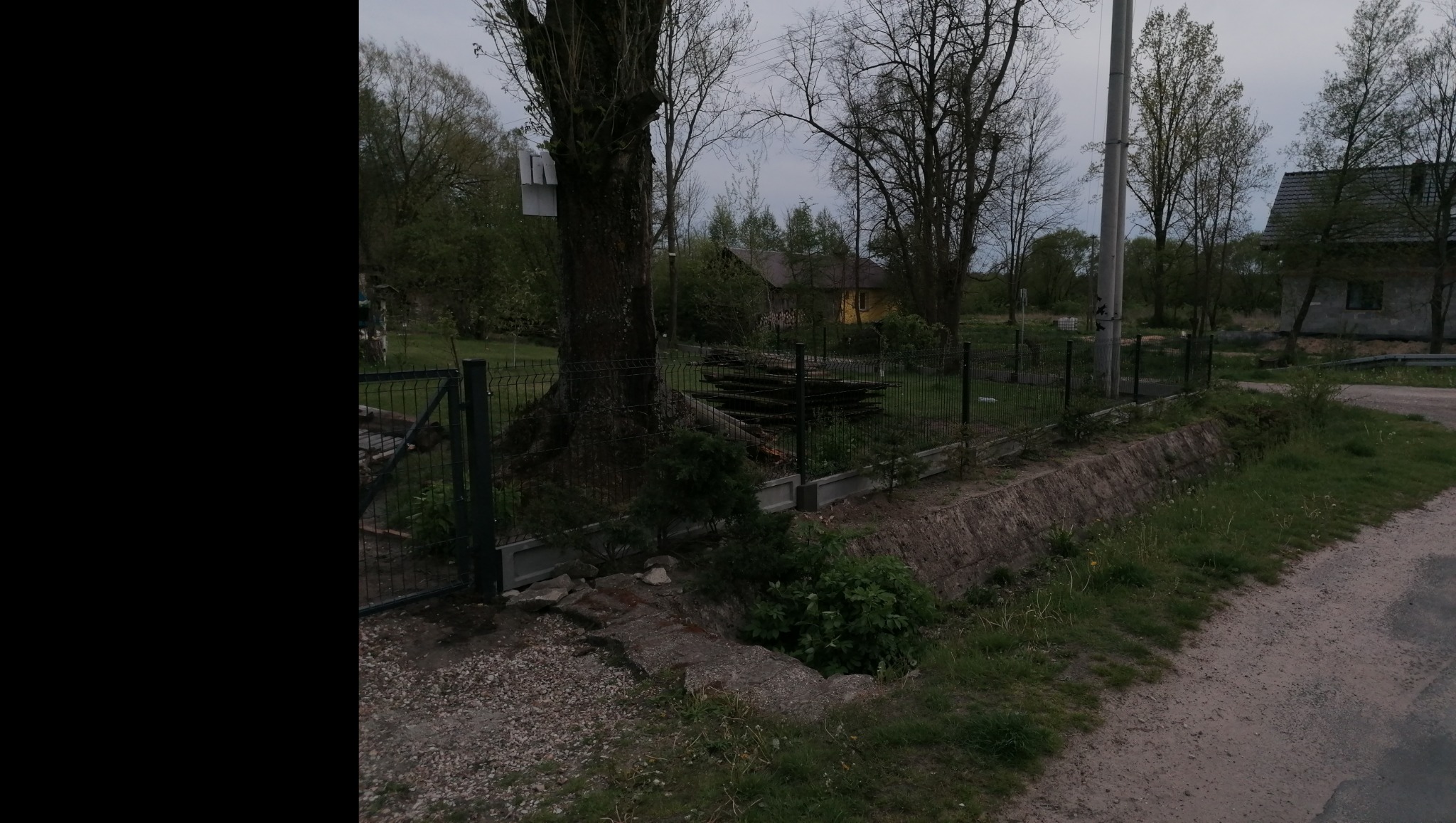 Metalowe ogrodzenie panelowe z podmurówką betonową, częściowo porośnięte roślinnością, obok drogi gruntowej, w tle domy i drzewa pod pochmurnym niebem.