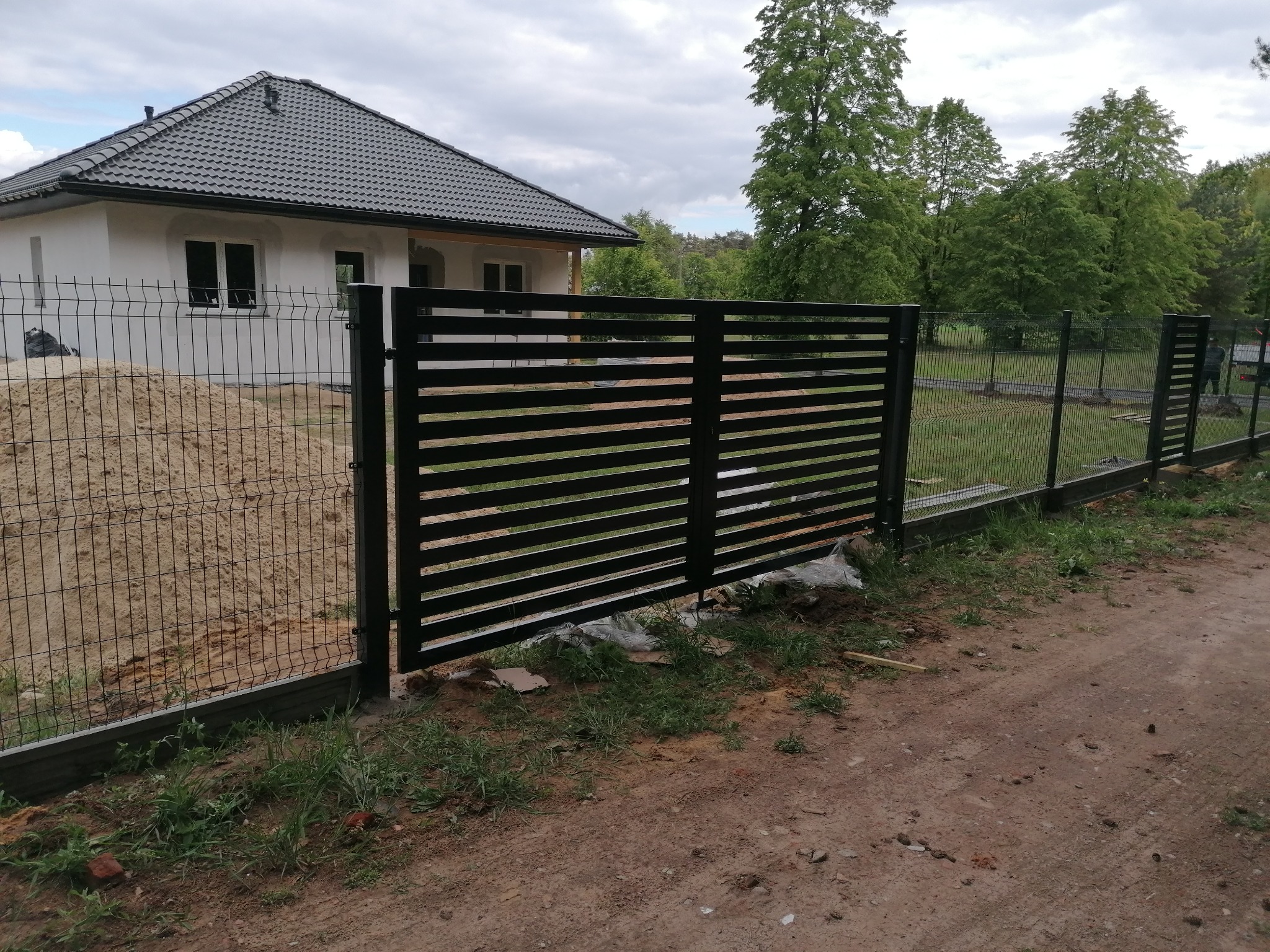 Nowoczesna, czarna brama przesuwna z poziomych profili, sąsiadująca z ogrodzeniem z siatki, na tle białego domu z grafitowym dachem i zieleni drzew.