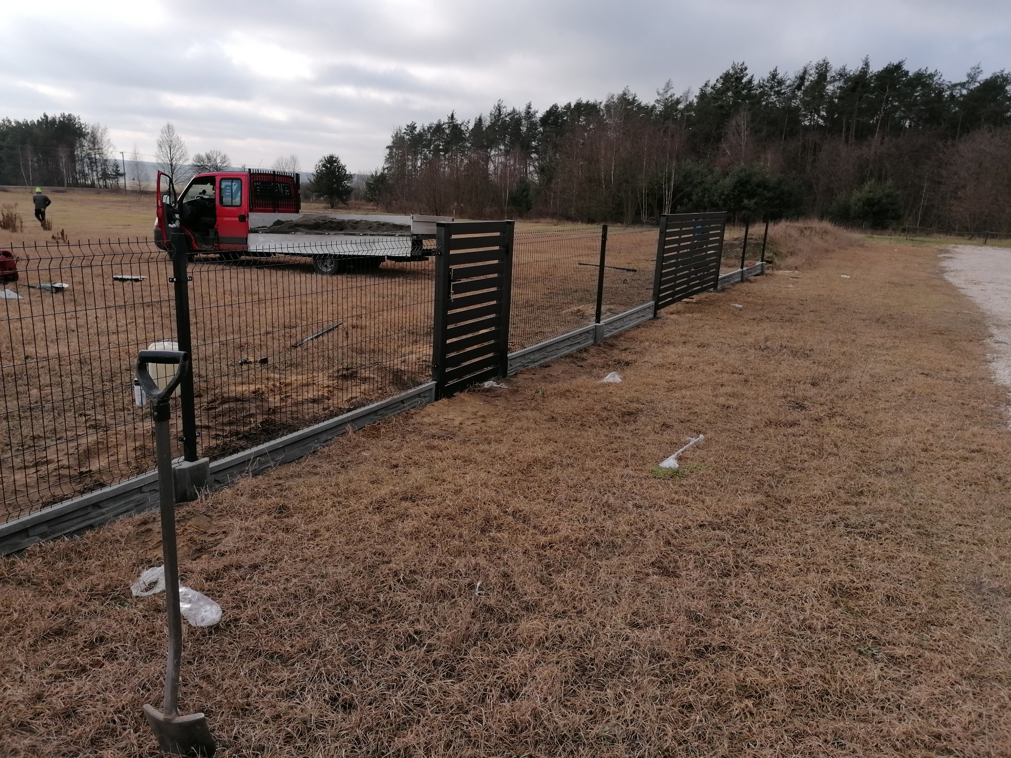 Fragment nowo montowanego ogrodzenia panelowego z podmurówką betonową, widoczny czerwony samochód dostawczy z przyczepą wypełnioną piaskiem, łopata wbita w ziemię, w tle las i pochmurne niebo.