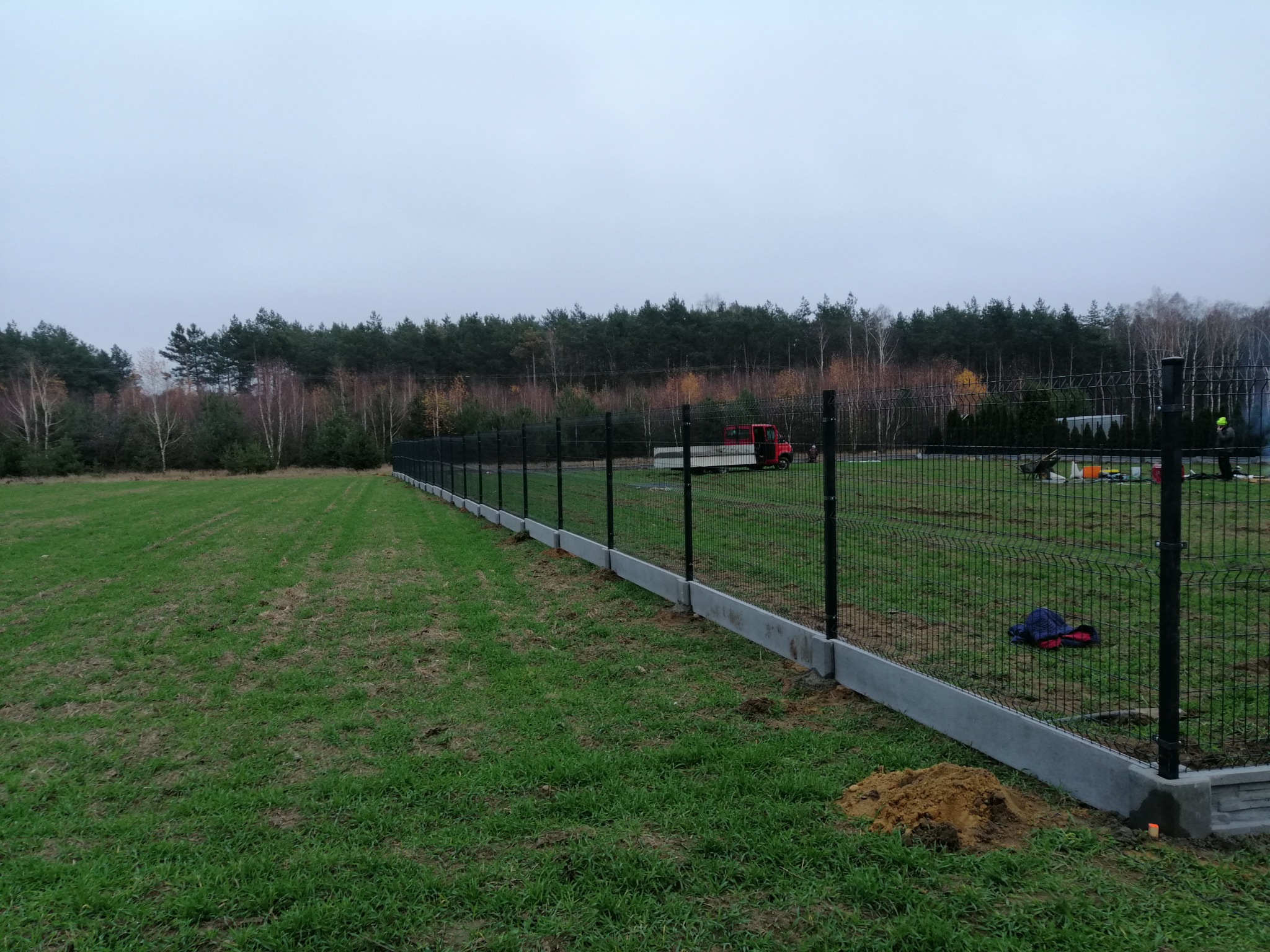 Długi fragment ogrodzenia panelowego z podmurówką betonową na skraju trawnika, w tle las i czerwony samochód dostawczy.