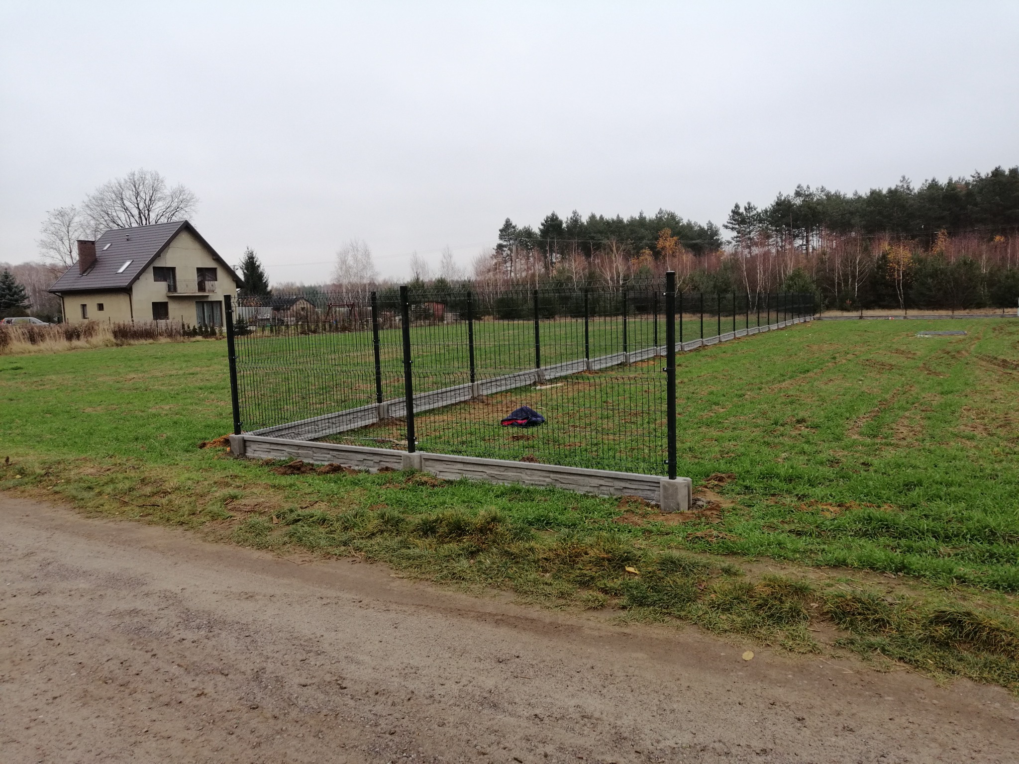 Częściowo zamontowane, panelowe ogrodzenie z podmurówką betonową na działce z domem w tle, widoczne ślady po pracach ziemnych.