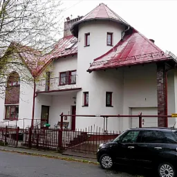 Biały dom z czerwoną dach&oacute;wką i wieżyczką, otoczony metalowym ogrodzeniem, z zaparkowanym czarnym samochodem na ulicy.