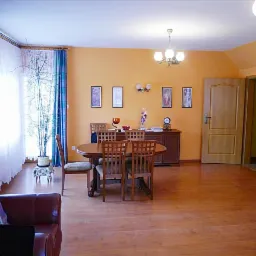 Jasne wnętrze salonu z drewnianą podłogą, okrągłym stołem jadalnym, krzesłami z tapicerką, pomarańczowymi ścianami i dekoracyjnymi zasłonami w oknie.