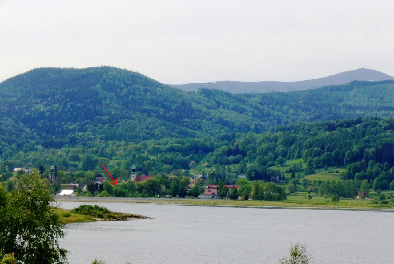 Pejzaż z widokiem na jezioro, zalesione wzgórza i budynki z wieżami w oddali, w tle góry.