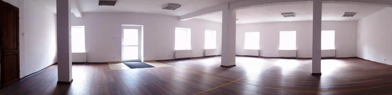 Panoramiczne ujęcie pustego, jasnego pomieszczenia z drewnianą podłogą, białymi ścianami, grzejnikami pod oknami i kolumnami nośnymi.