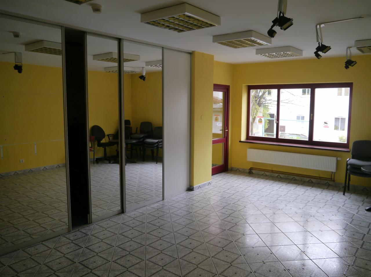 Pusty pokój biurowy z żółtymi ścianami, szarą podłogą w romby, lustrzaną szafą wnękową, oknem z widokiem na budynek i lampami sufitowymi