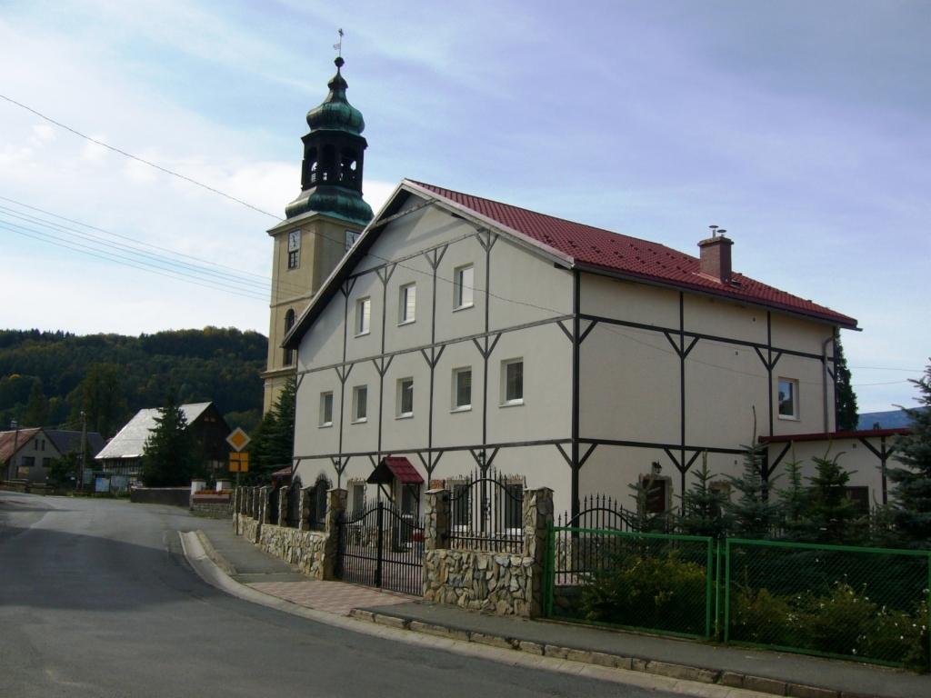 Dwupiętrowy budynek z czerwonym dachem i czarnymi belkami dekoracyjnymi, widoczny z ulicy w małej miejscowości, z wieżą kościelną w tle.