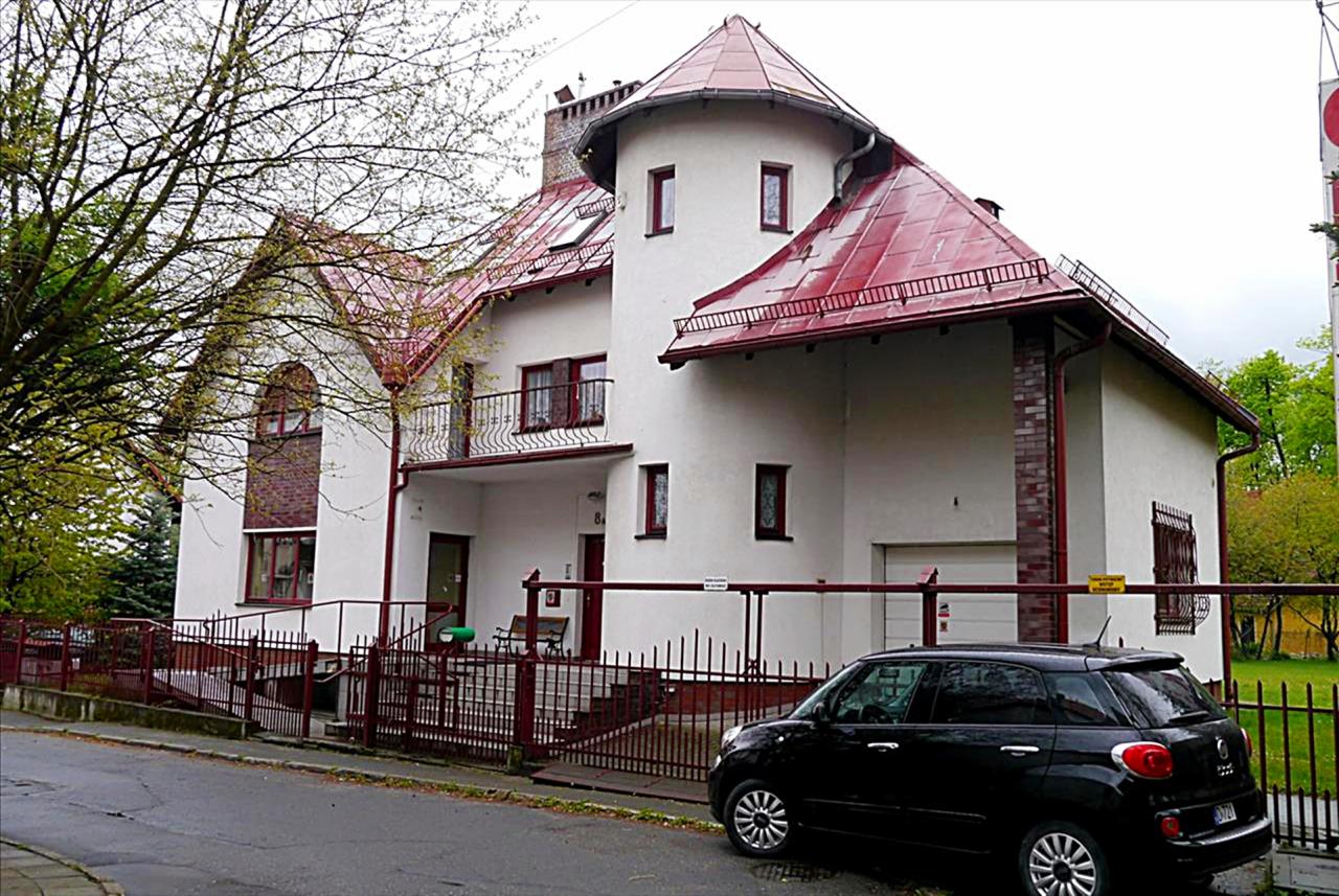 Biały dom z czerwoną dachówką i wieżyczką, otoczony metalowym ogrodzeniem, z zaparkowanym czarnym samochodem na ulicy.