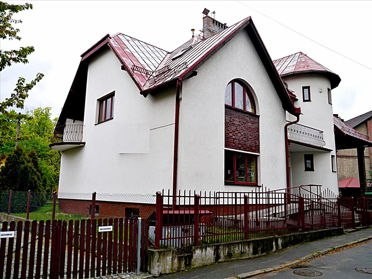 Dwupiętrowy dom z wieżyczką, białymi ścianami, czerwoną dachówką i drewnianym ogrodzeniem, widziany z poziomu ulicy.
