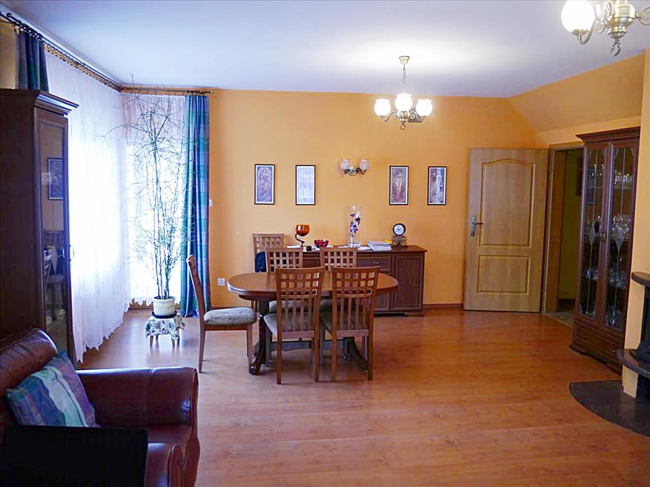 Jasne wnętrze salonu z drewnianą podłogą, okrągłym stołem jadalnym, krzesłami z tapicerką, pomarańczowymi ścianami i dekoracyjnymi zasłonami w oknie.