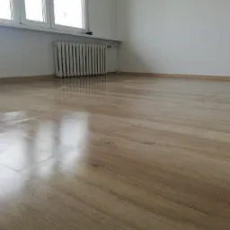 Jasne panele podłogowe w pokoju po ułożeniu, widoczne delikatne ślady czyszczenia, białe ściany, grzejnik pod oknem.