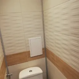 Wykończona toaleta z beżowymi płytkami imitującymi drewno, białym kompaktem WC marki Cersanit i ukrytym dostępem do instalacji za pomocą białej klapy rewizyjnej.