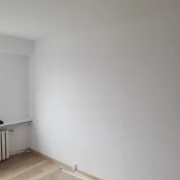 Pomalowane na biało wnętrze pokoju z jasną podłogą panelową, widoczne listwy przypodłogowe i wystające kable ze stropu.