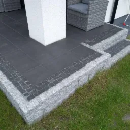 Nowoczesny taras z ciemnoszarą płytką i obrzeżem z jasnoszarego granitu, schody wejściowe z kostki granitowej, widok z poziomu trawnika.