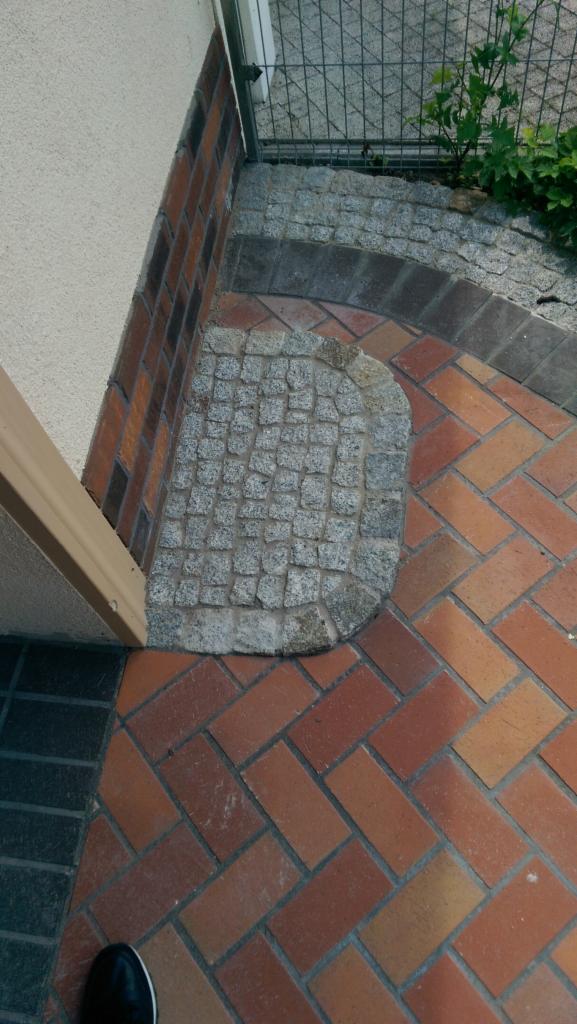 Ułożona z cegły klinkierowej ścieżka w jodełkę, z wykończeniem z kostki granitowej przy wejściu do budynku, otoczona murem z cegły i siatką ogrodzeniową.