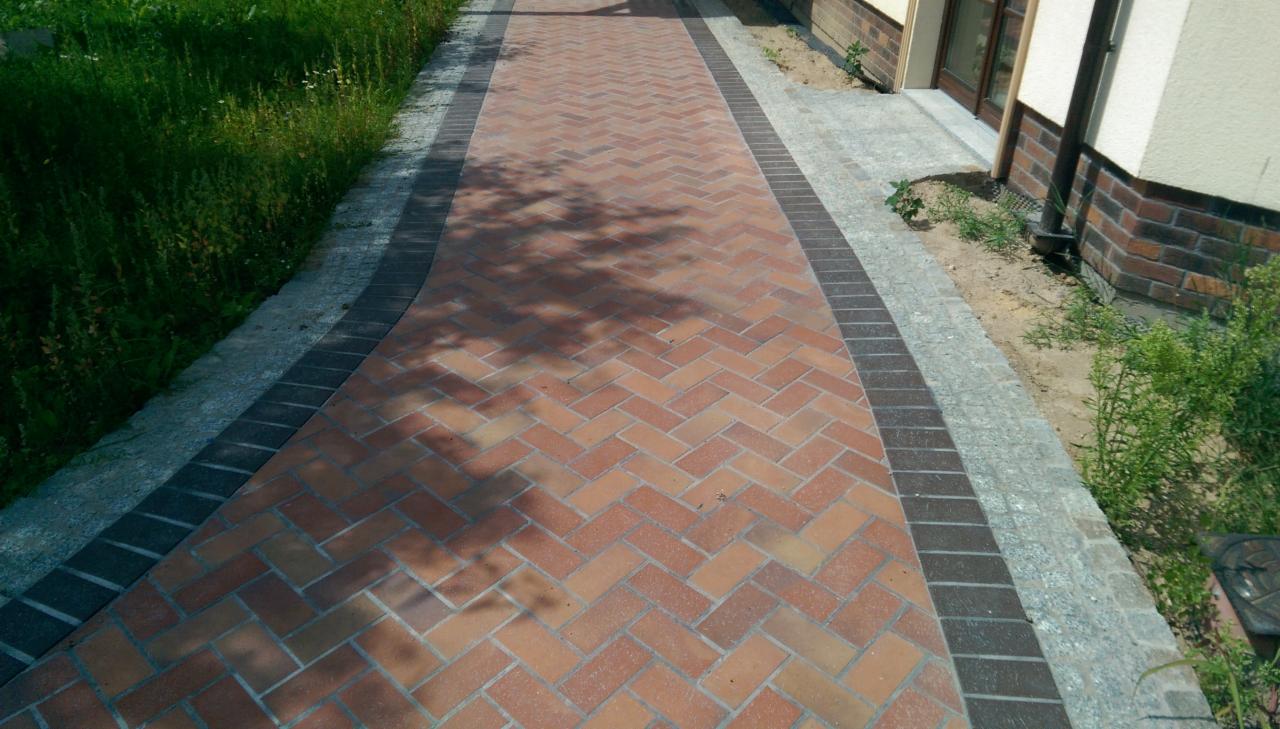 Ceglana ścieżka ułożona w jodełkę z ciemniejszym obramowaniem, prowadząca wzdłuż budynku z ceglaną podmurówką, z widoczną zielenią po bokach.