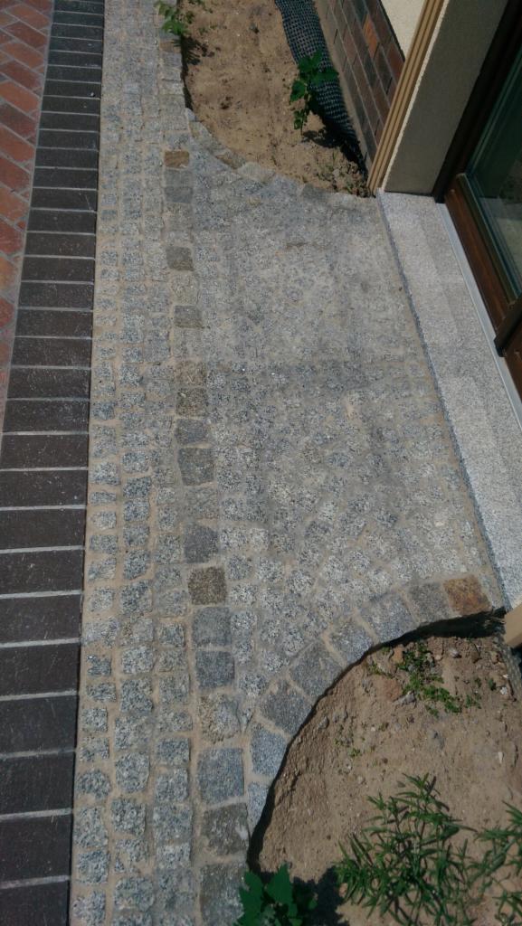 Granitowa kostka brukowa ułożona wzdłuż ścieżki i przylegająca do budynku z widocznym oknem, z fragmentami ziemi i zieleni po bokach.