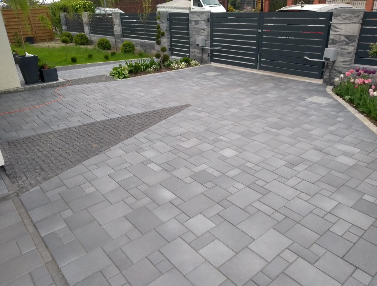 Nowoczesny podjazd z szarej kostki brukowej ułożonej we wzór, z elementami z ciemniejszej kostki, brama wjazdowa z automatyką, zadbana zieleń w tle.