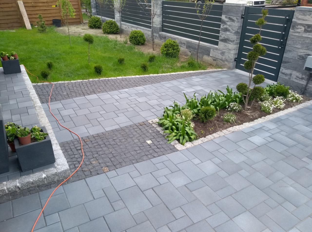 Szara kostka brukowa ułożona w geometryczny wzór z elementami z ciemniejszej kostki i rabatami kwiatowymi, otoczona trawnikiem i nowoczesnym ogrodzeniem.