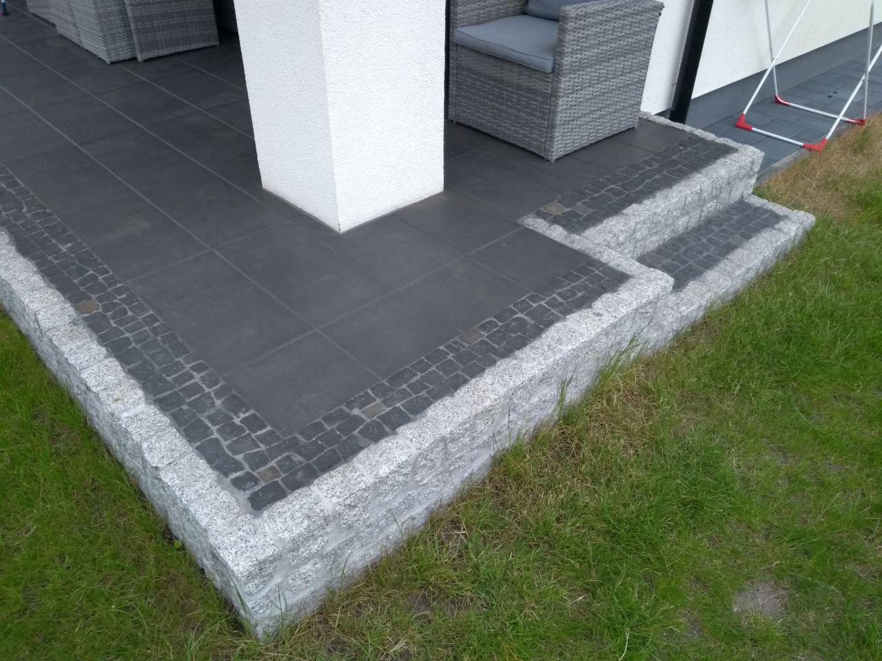 Nowoczesny taras z ciemnoszarą płytką i obrzeżem z jasnoszarego granitu, schody wejściowe z kostki granitowej, widok z poziomu trawnika.