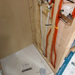 Montaż brodzika w kabinie prysznicowej z widocznymi instalacjami wodno-kanalizacyjnymi i elektrycznymi w ścianie z boazerii, częściowo wykończonej panelami podłogowymi Krono Original.