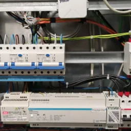 Rozdzielnica elektryczna z zainstalowanymi bezpiecznikami, modułami sterującymi i przewodami o różnych kolorach, widok z bliska na elementy instalacji elektrycznej w metalowej obudowie.