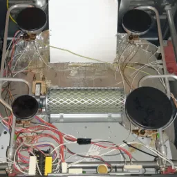 Wnętrze rozmontowanej kuchenki gazowej z widocznymi palnikami, przewodami i elementami grzejnymi, ukazujące skomplikowaną budowę urządzenia.