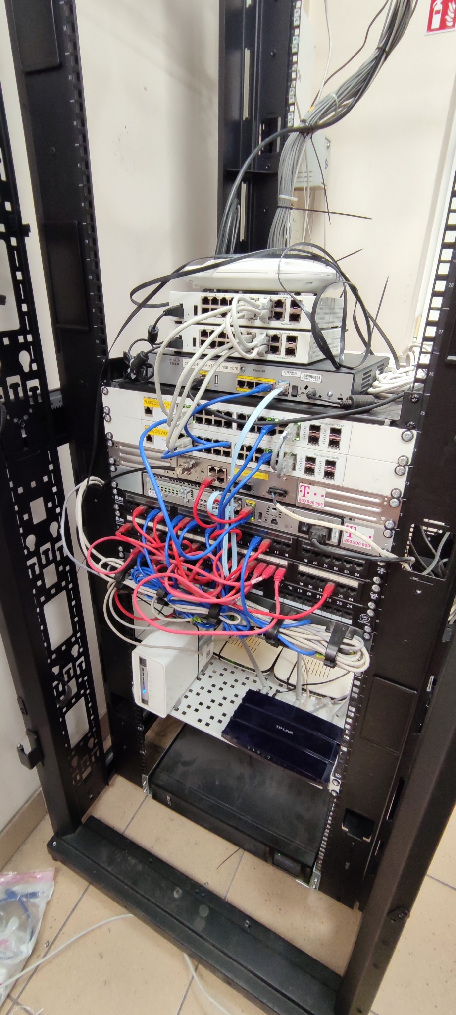 Szafa rack z zainstalowanymi urządzeniami sieciowymi: routery Cisco, przełączniki, patch panele, okablowanie strukturalne w kolorach czerwonym, niebieskim i białym, zasilacze UPS na perforowanej...