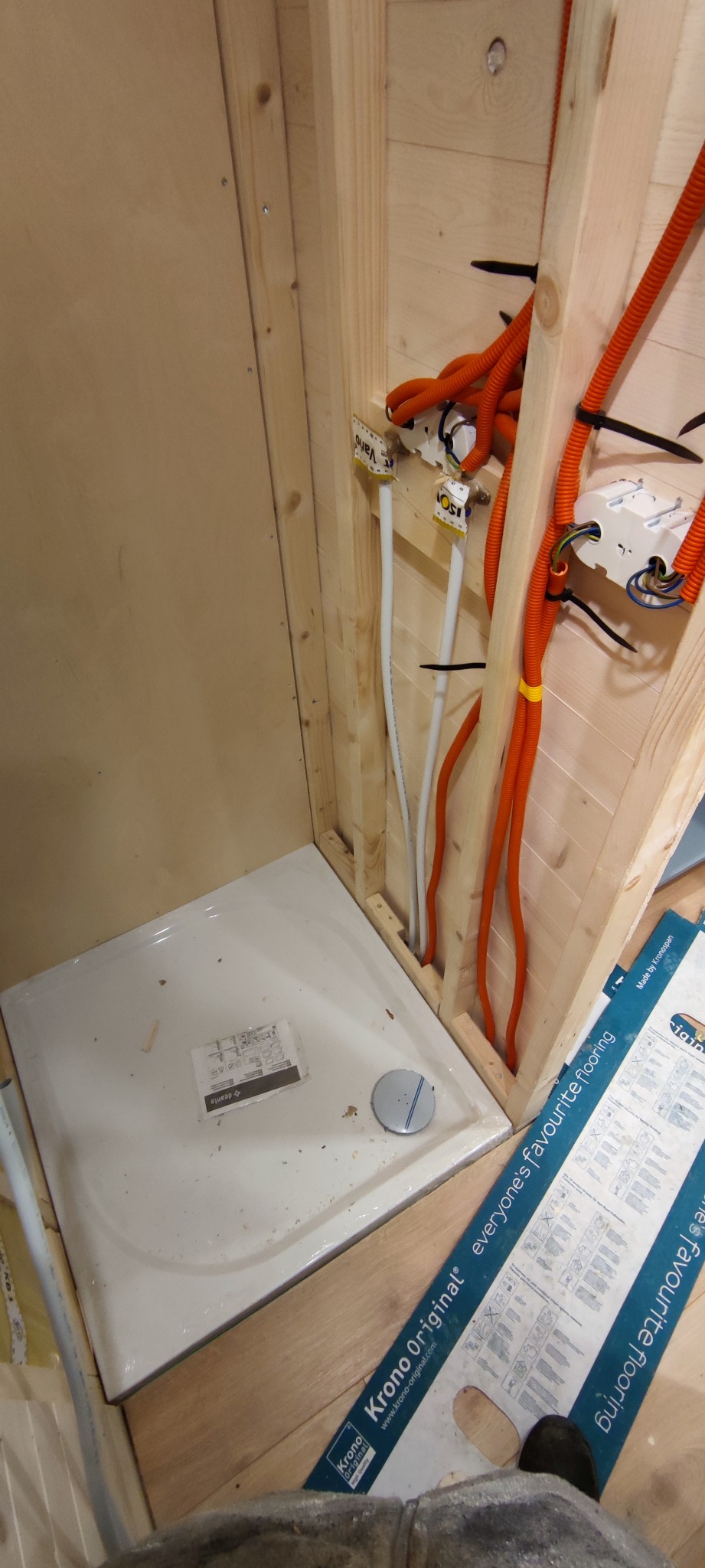 Montaż brodzika w kabinie prysznicowej z widocznymi instalacjami wodno-kanalizacyjnymi i elektrycznymi w ścianie z boazerii, częściowo wykończonej panelami podłogowymi Krono Original.