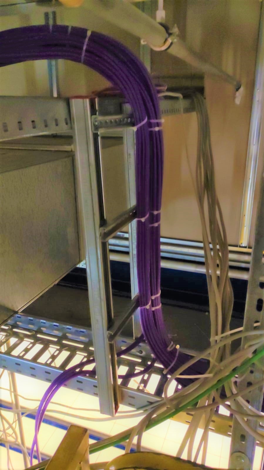 Pęknięcie fioletowych kabli spiętych opaskami, prowadzonych w metalowej konstrukcji pod sufitem, widok z dołu.