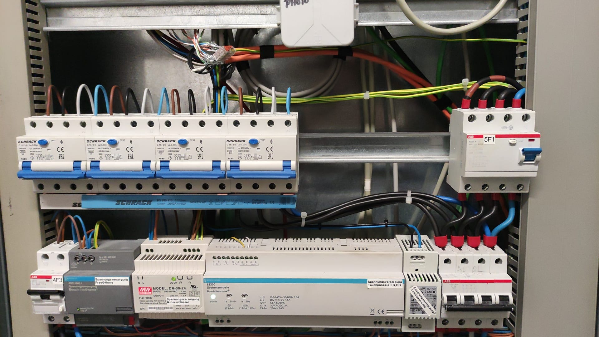 Rozdzielnica elektryczna z zainstalowanymi bezpiecznikami, modułami sterującymi i przewodami o różnych kolorach, widok z bliska na elementy instalacji elektrycznej w metalowej obudowie.