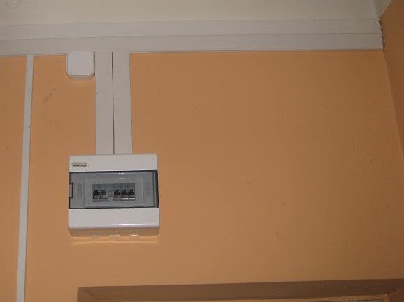 Zamknięta, biała skrzynka rozdzielcza z szarą, przezroczystą pokrywą, zamontowana na pomarańczowej ścianie, z białą listwą przypodłogową u góry i białymi kablami w listwach biegnącymi do góry.