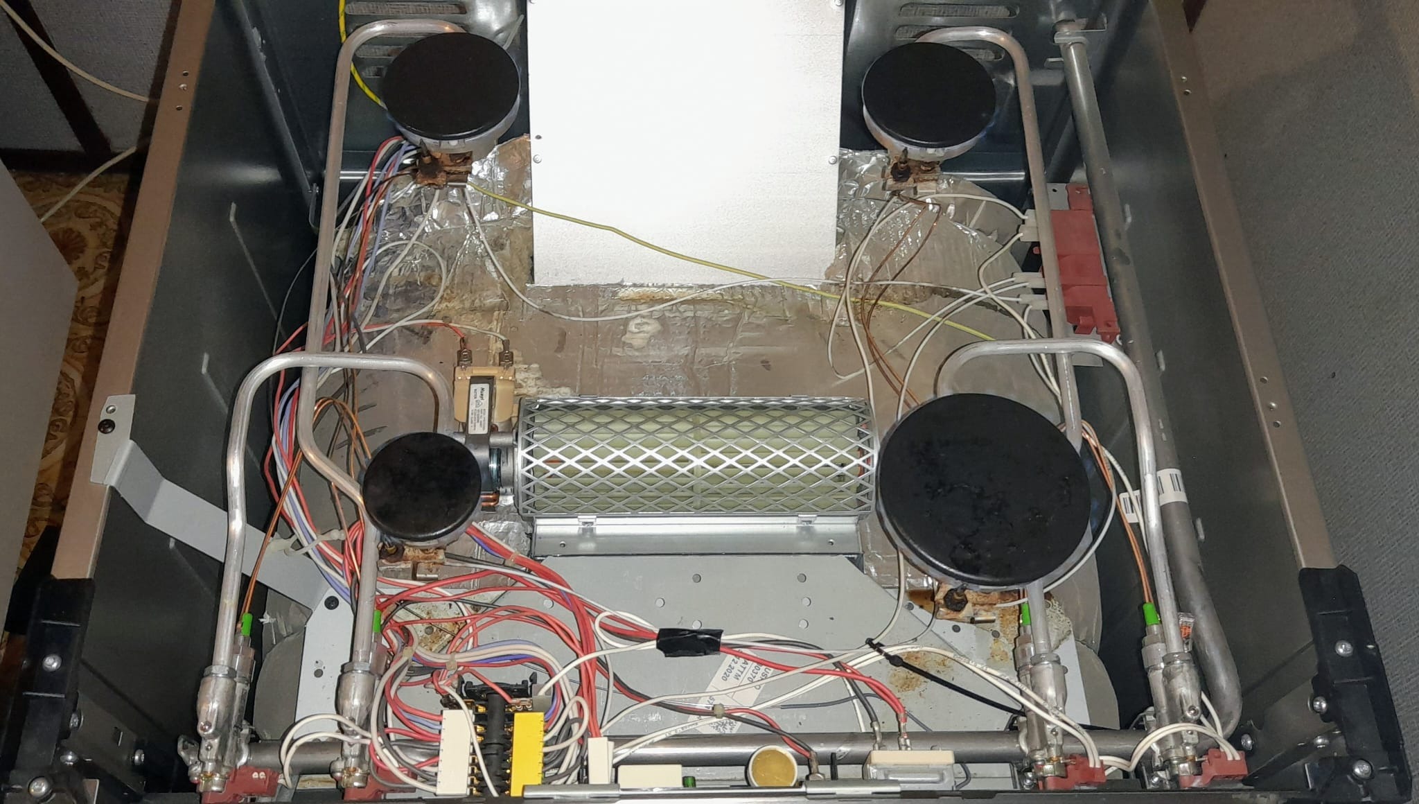 Wnętrze rozmontowanej kuchenki gazowej z widocznymi palnikami, przewodami i elementami grzejnymi, ukazujące skomplikowaną budowę urządzenia.