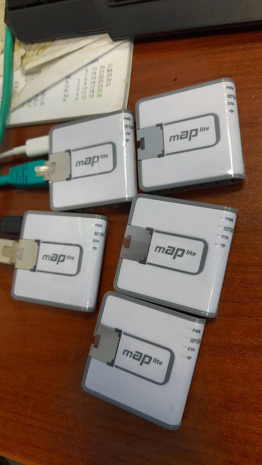 Cztery białe routery Map Lite z podłączonymi kablami Ethernet leżące na drewnianym biurku, obok kalendarza.
