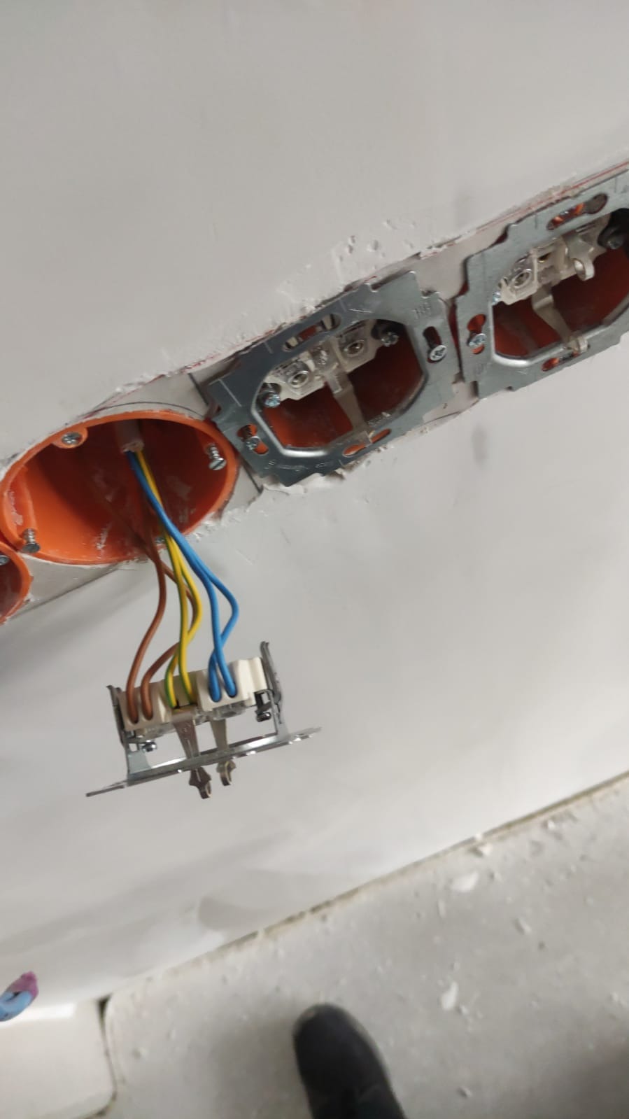 Montaż gniazdek elektrycznych w puszce podtynkowej, widoczne przewody: niebieski, żółty, brązowy, oraz metalowe elementy mocujące.