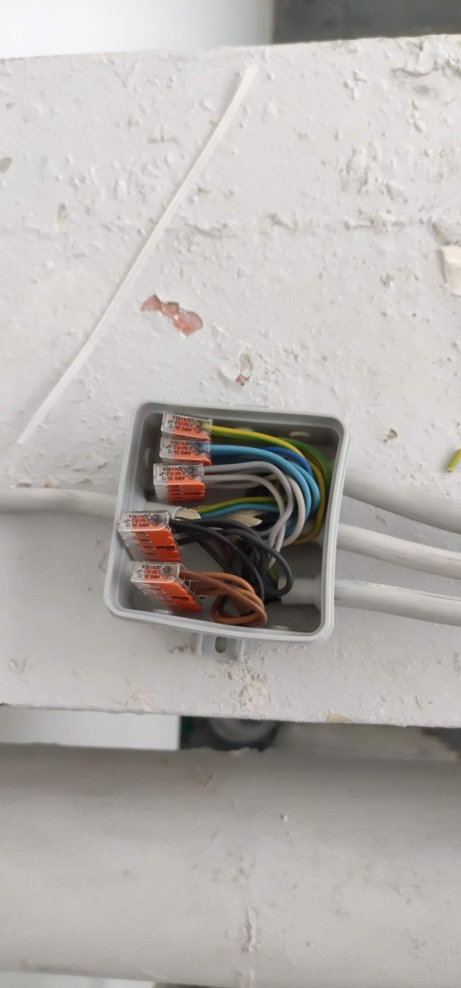 Otwarta puszka elektryczna z widocznymi przewodami w różnych kolorach, połączonymi za pomocą złączek Wago, umieszczona na pomalowanej, nierównej powierzchni.