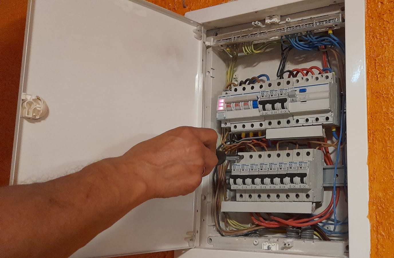 Otwarta skrzynka elektryczna z bezpiecznikami, w której widoczna dłoń z próbnikiem sprawdza obwód elektryczny.