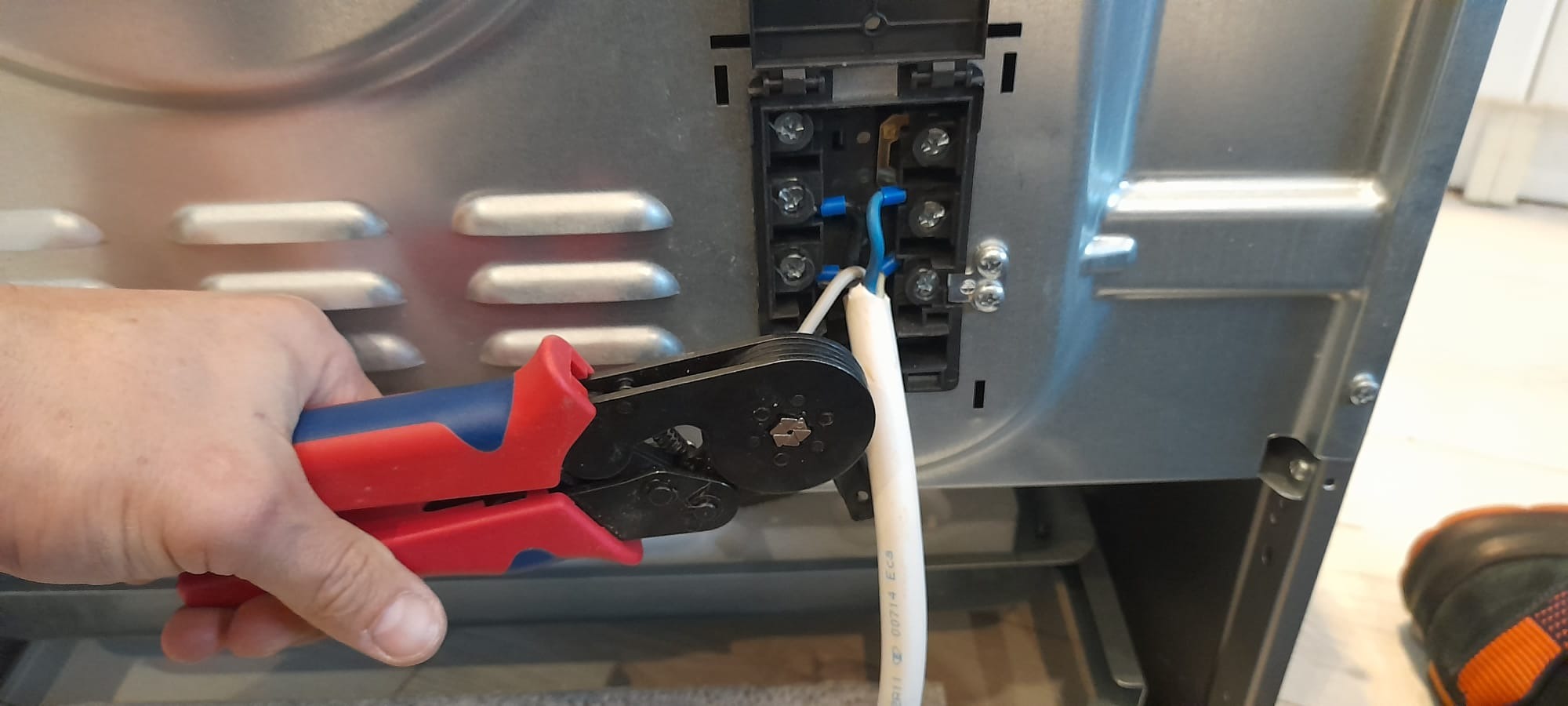 Podłączenie przewodu elektrycznego do piekarnika za pomocą zaciskarki z czerwono-niebieską rękojeścią, widoczna tylna ściana piekarnika z przyłączem elektrycznym.