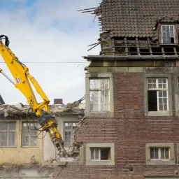 Żółty koparko-ładowarka z hydraulicznym młotem kruszy ceglany budynek z odsłoniętą konstrukcją dachu i widocznymi oknami, obok widoczna elewacja sąsiedniego budynku w trakcie prac.