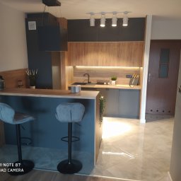 Maryka Ryszard Kapałka - Nowoczesna kuchnia z drewnianymi frontami szafek górnych i ciemnoszarymi dolnymi, wyspą z dwoma szarymi hokerami i oświetleniem punktowym.